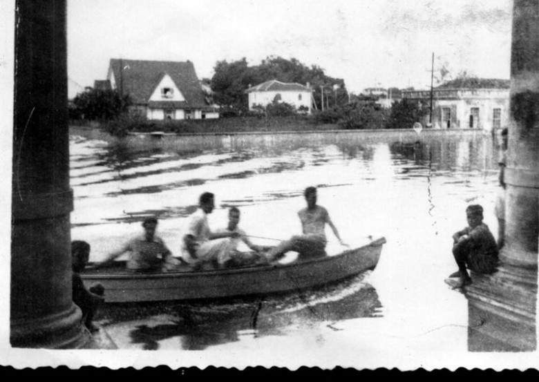 Na foto antiga vemos moradores passando na frente da estação com um pequeno barco. 